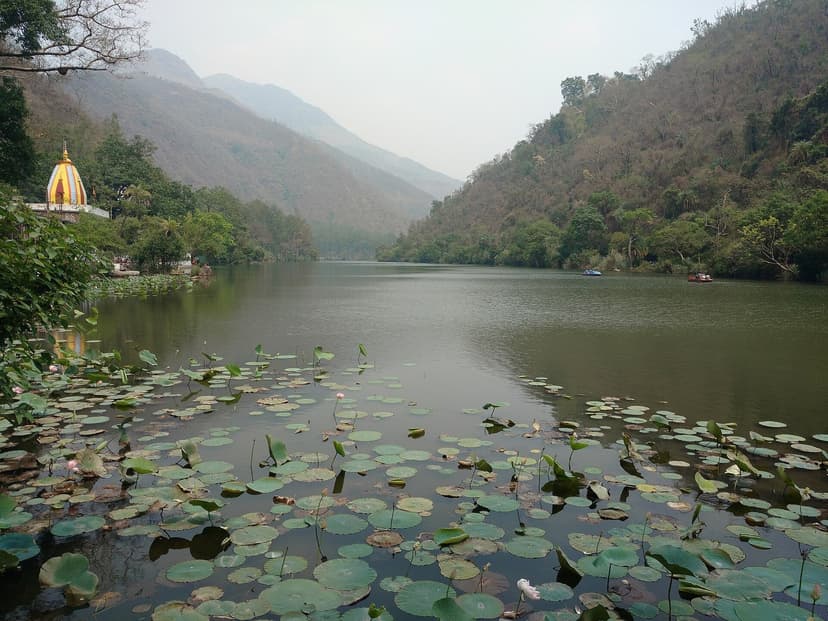 Renuka Ji Lake in Sirmaur district, still water surface with lily pads, a small red-and-yellow shrine visible on the left bank, forested hills on both sides and a sliver of temple complex in the middle distance