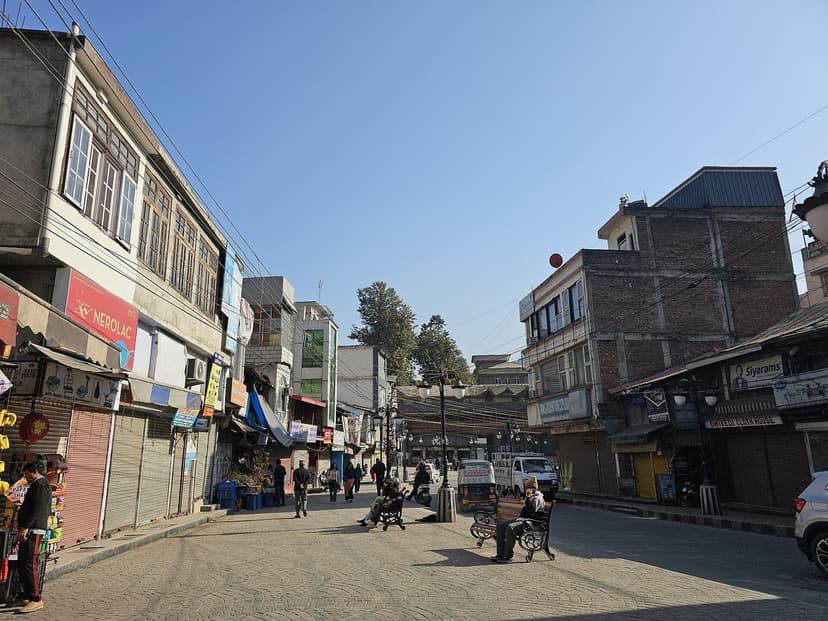A central street in Mandi town, Himachal Pradesh — paved square with shops, low-rise buildings and a few people walking, the small district town where Mandyali Dham is the standard Boti repertoire and Mandi Shivratri brings the largest community feasts of the year