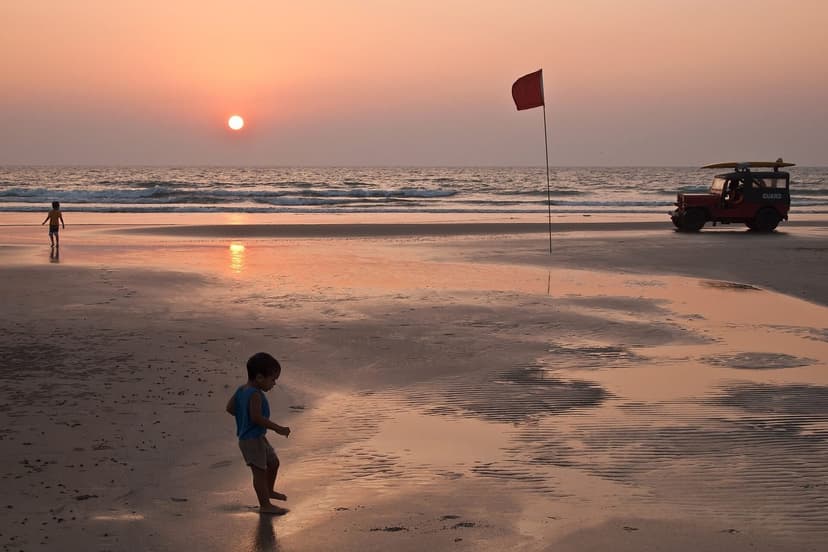 A near-empty Mandrem beach at sunset with a red lifeguard flag and a lifeguard jeep on the wet sand, a child walking in the foreground