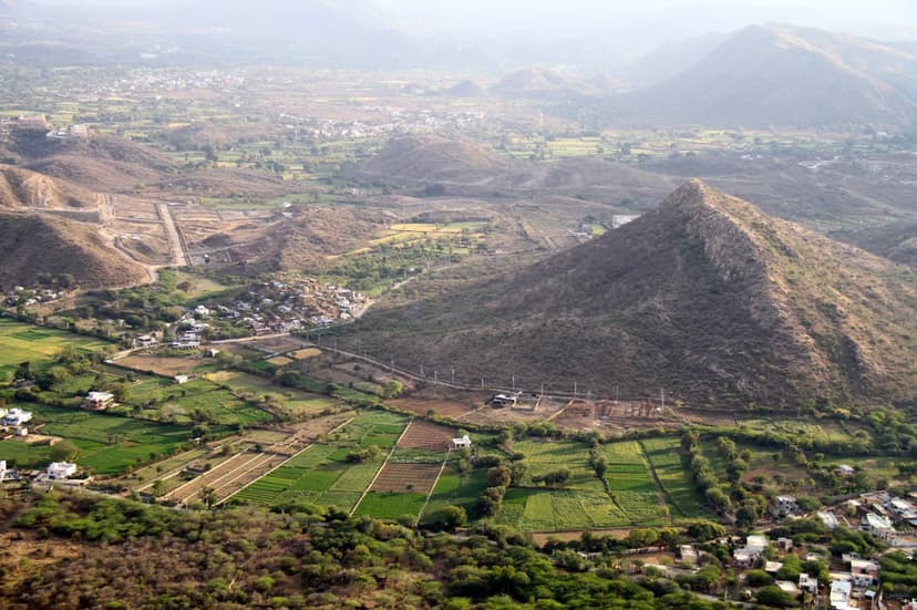 Elevated view over a patchwork of green farm fields and scattered village houses in the countryside of southern Rajasthan, Aravalli hills behind.