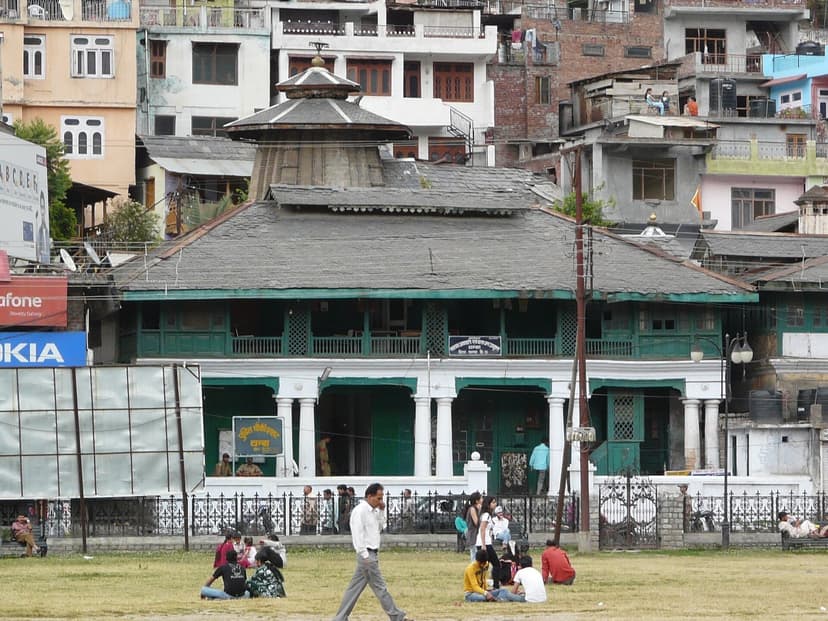 Old green-and-white civic building at the edge of the Chaugan in Chamba with the shikhara of Champavati temple rising behind and people sitting on the grass