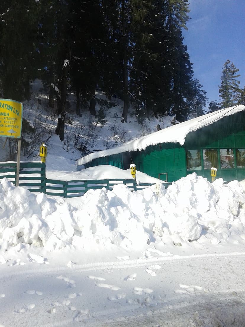 A green HPTDC forest cabin at Narkanda buried under deep fresh snow, with snow piled on the roof and fenced boundary
