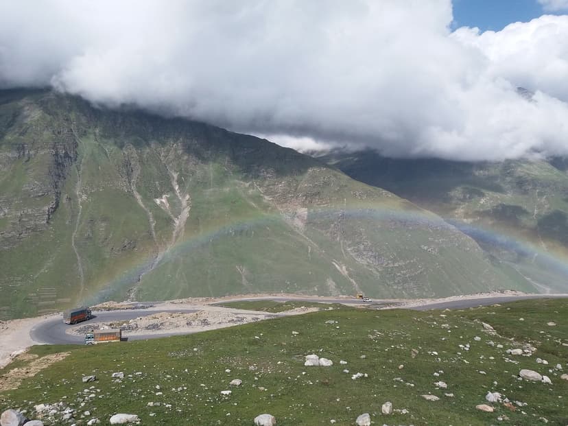 Hairpin bends of the old Rohtang Pass road carved into a green Himalayan slope, with a full rainbow arching across the valley after a passing rain shower and clouds clinging to the ridge above