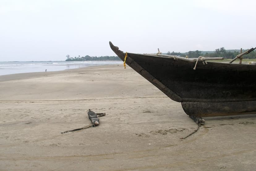 A wide, near-empty Morjim beach in North Goa with a traditional wooden fishing boat pulled up on the sand and a palm-lined headland behind