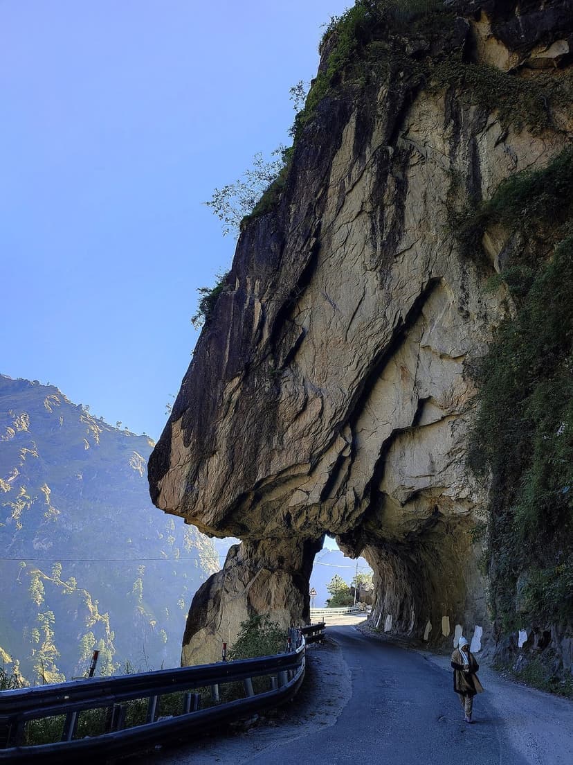 Massive rock overhang on the Kinnaur highway at Taranda cliff, with the road carved directly under an enormous jutting rock face and a person walking for scale