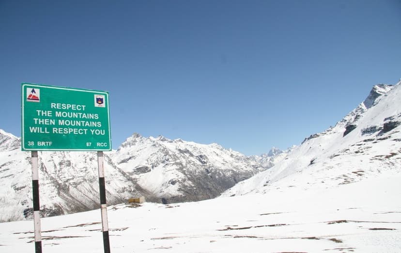 Green BRO road sign at Rohtang Pass summit reading Respect The Mountains Then Mountains Will Respect You, with snow-covered landscape and peaks behind it, marked 38 BRTF 87 RCC