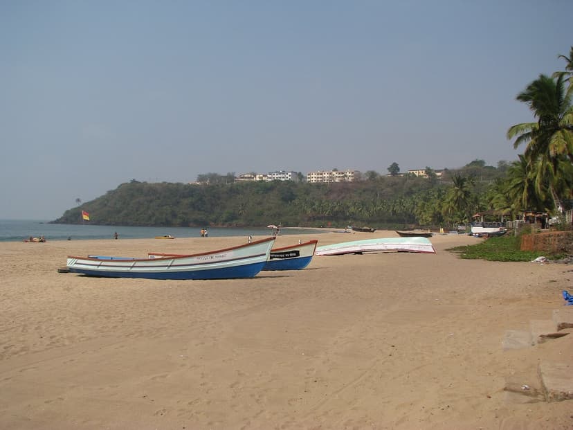 Fishing boats pulled up on the sand at Bogmalo beach in south Goa, with a green headland and hillside buildings behind and a calm sea under a blue sky.