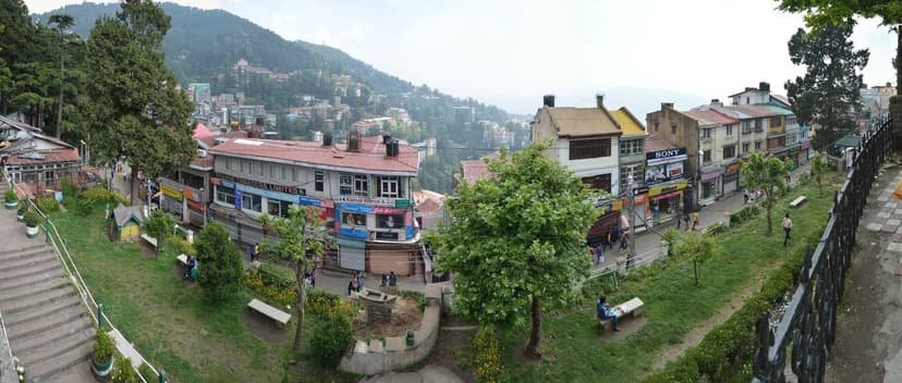 A panoramic view of the Ridge and Mall Road in Shimla — green slopes, deodar trees and red-tile roofs of colonial-era buildings, Mall Road shopfronts including Sony and other signs, pedestrians on the promenade