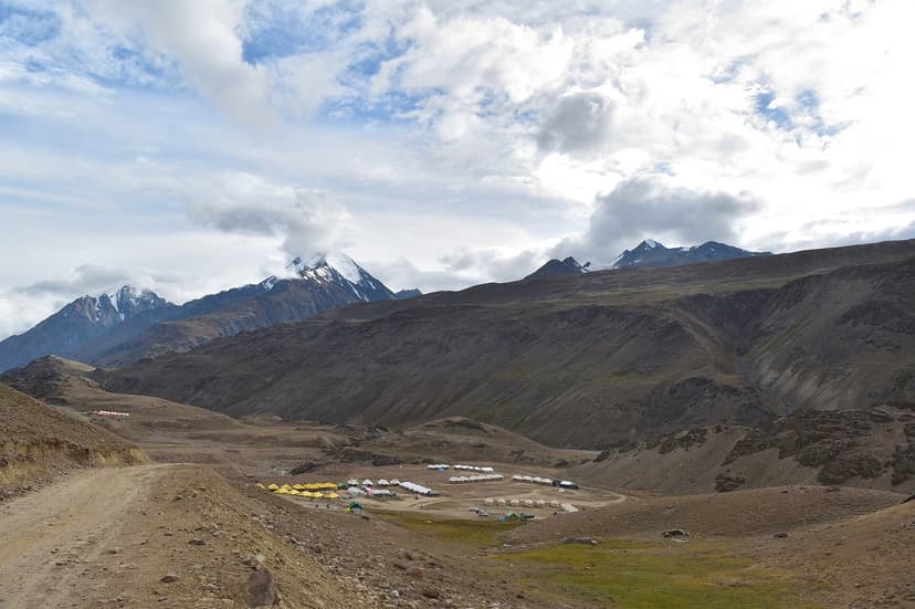Wide bowl of brown moraine and green meadow at Chandratal Lake, with a yellow-roofed campsite cluster in the middle distance and snow peaks rising along the horizon under a part-cloudy sky