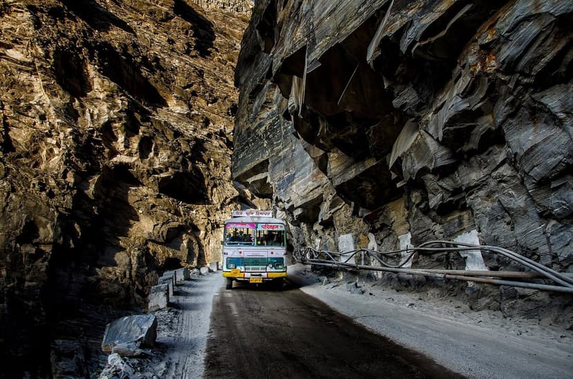 A colourful bus squeezing through a narrow rock tunnel carved into the cliff face at Taranda Dhank on the Kinnaur highway, with bare rock walls towering on both sides