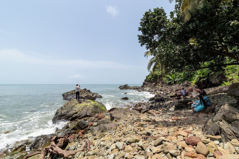 A rocky pebble cove below a forested headland at Cabo de Rama, with a person standing on a boulder photographing the sea