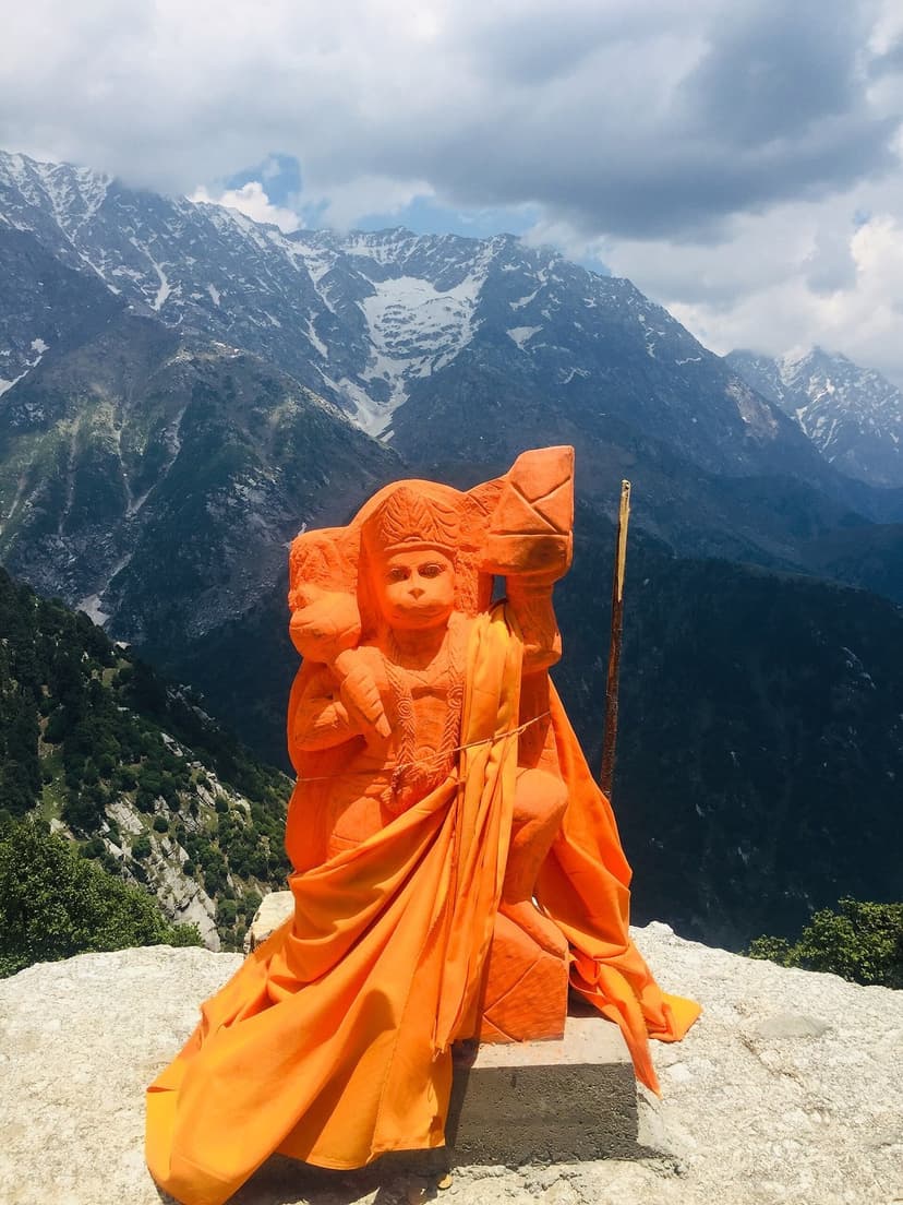 Saffron-draped Hanuman statue on the Triund ridge with snow-capped Dhauladhar peaks rising behind and pine valley below