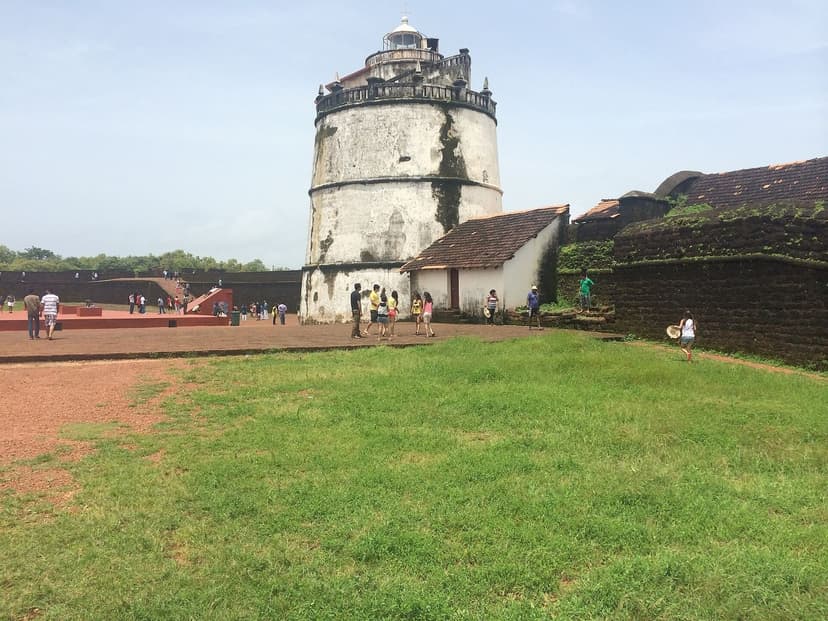 The round white lighthouse tower and laterite walls of the seventeenth-century Fort Aguada in North Goa, on a green lawn under a bright sky, a few distant visitors.