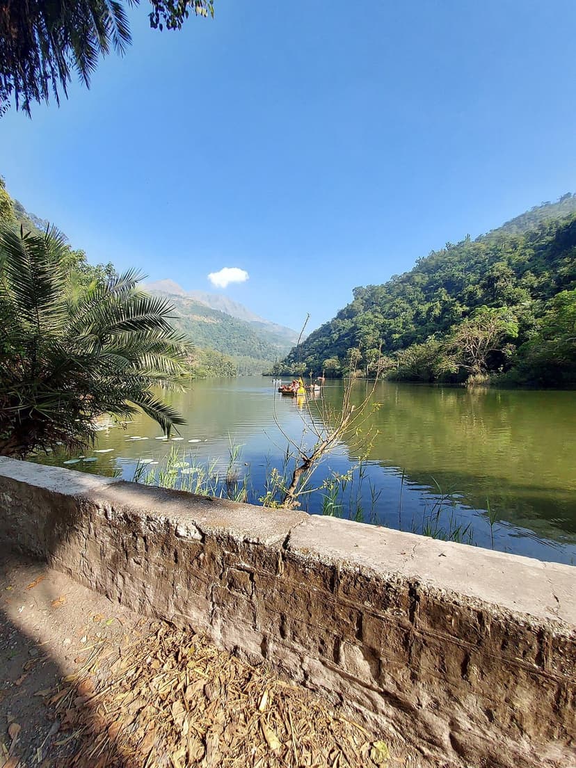 Renuka Lake in Sirmaur district — a calm green lake shaped like a reclining woman, surrounded by forested hills, a small boat in the middle distance, with a stone wall along a path in the foreground