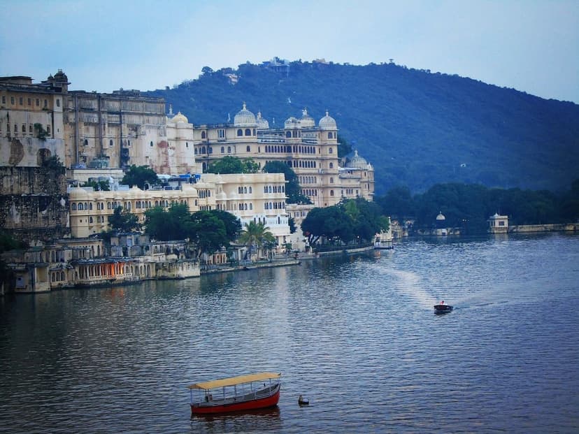 The City Palace complex and old-city buildings of Udaipur rising from Lake Pichola at dusk, tourist boats on the water and hills behind.