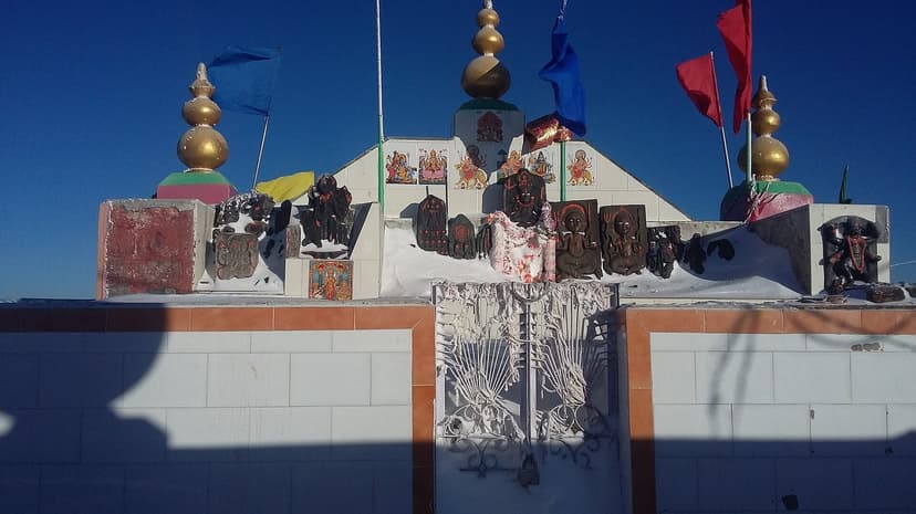 The roofless Shikari Devi temple in winter, a line of dark stone deity images standing in the open under a deep blue sky with snow and prayer flags