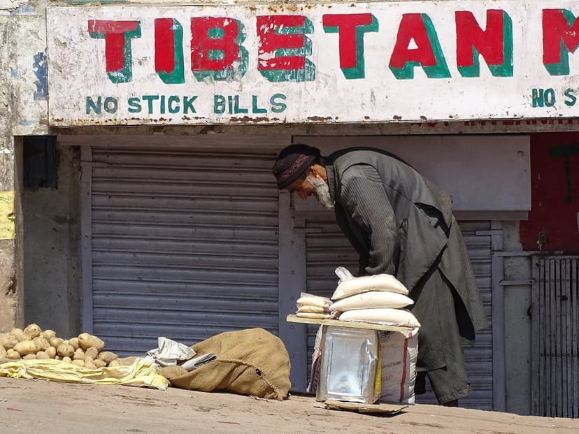 Elderly Tibetan vendor outside a shuttered shop with Tibetan-script signage in McLeod Ganj