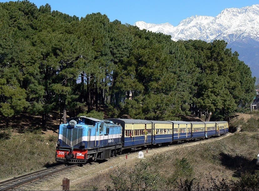 A blue-and-yellow Kangra Valley Railway narrow-gauge train on the track with snow-capped Dhauladhar peaks rising behind