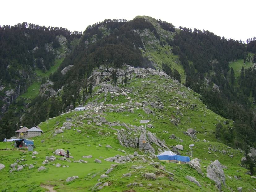 Trail going upward through a steep boulder-strewn green hillside with a small shelter visible