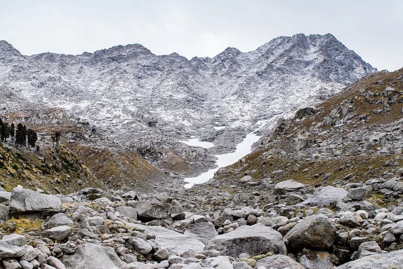 Steep boulder field with snow-dusted Dhauladhar peaks above, looking up toward Indrahar Pass